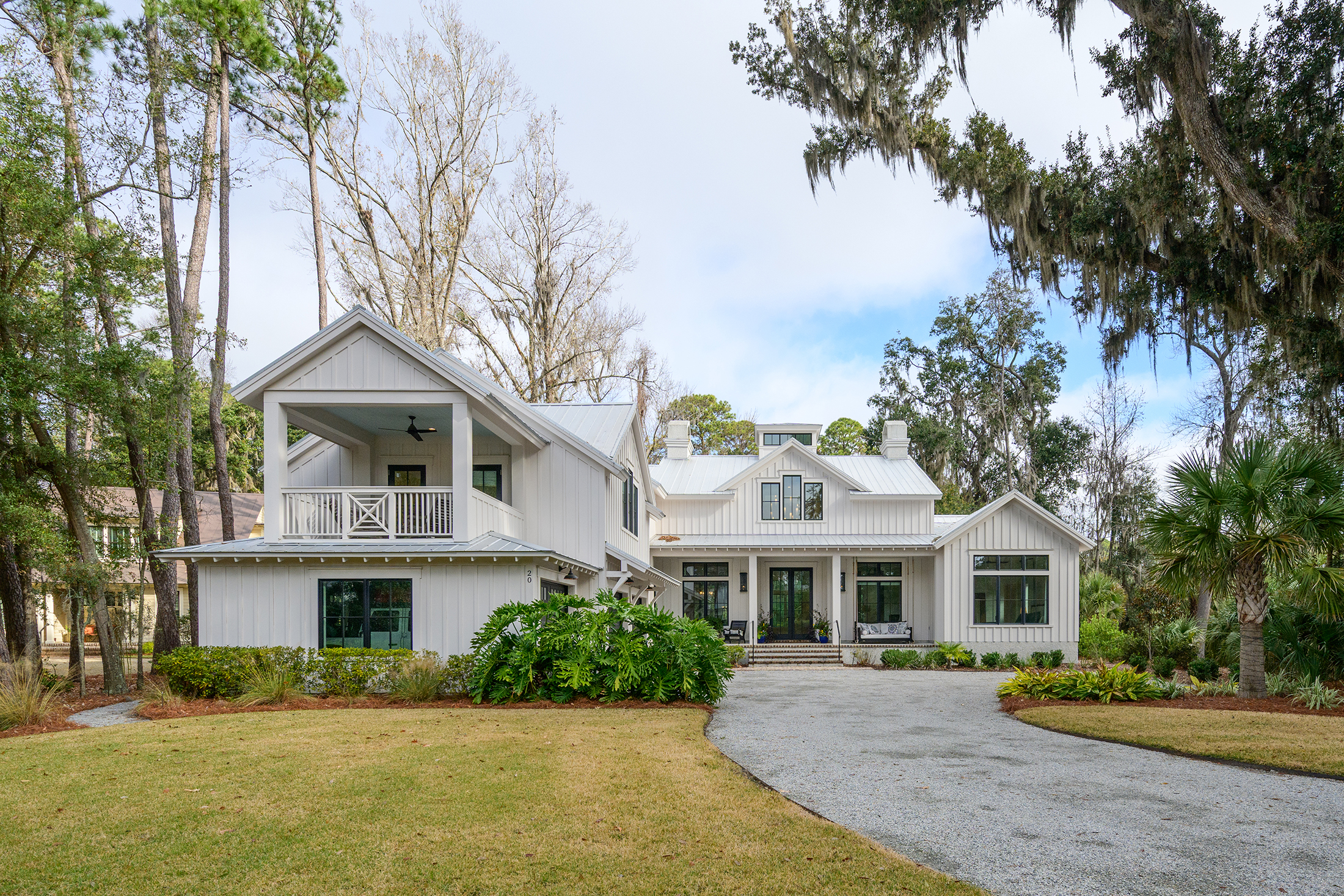 A large, modern farmhouse-style home with white siding and a metal roof, featuring a covered balcony and front porch, surrounded by trees and landscaped greenery—a true A-list retreat set to inspire in February 2026.