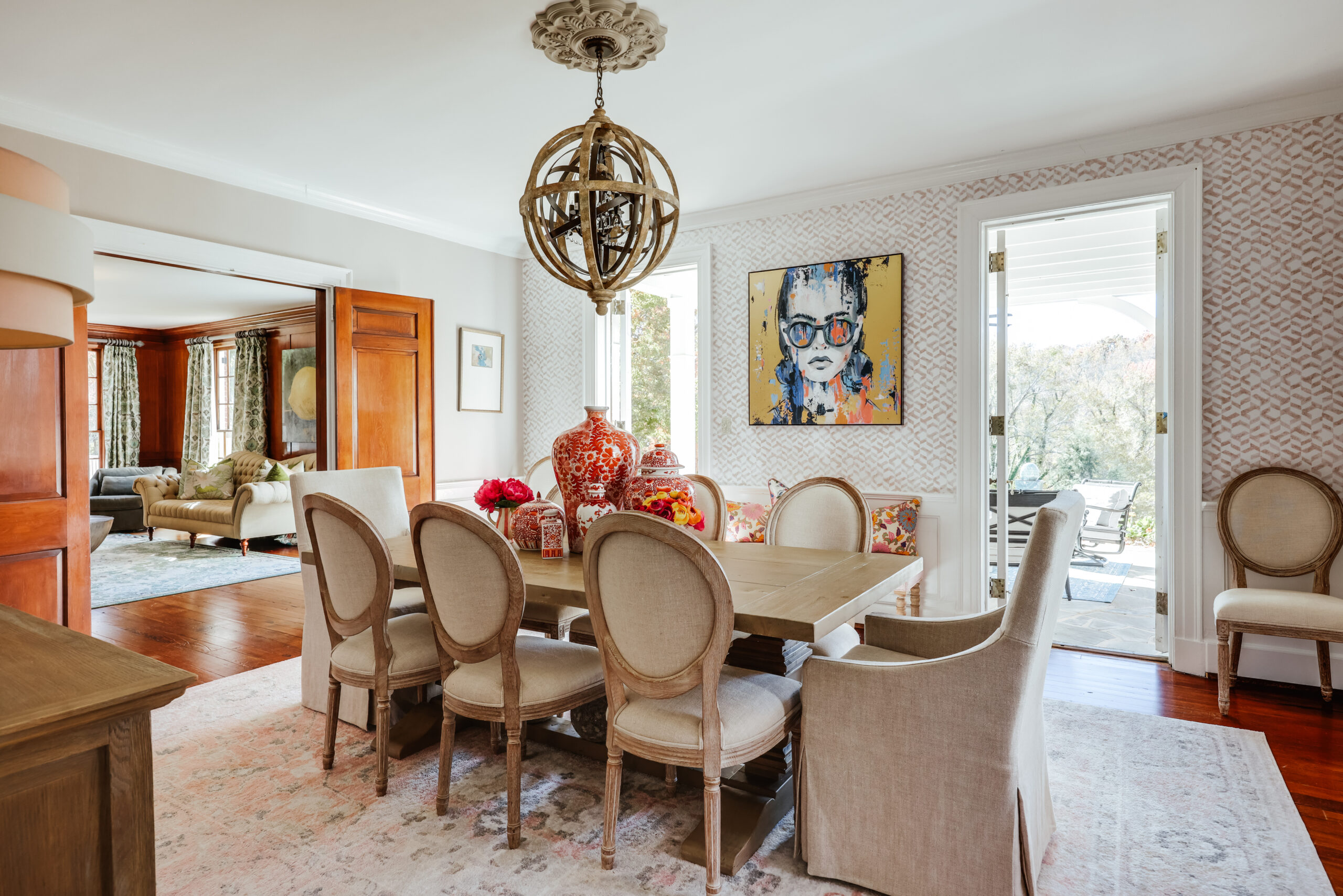 A bright dining room with a wooden table, eight upholstered chairs, and a modern chandelier. Colorful vases and flowers sit on the table, while an A-list: February 2026 portrait adds vibrance to patterned wallpaper near glass doors to a patio.