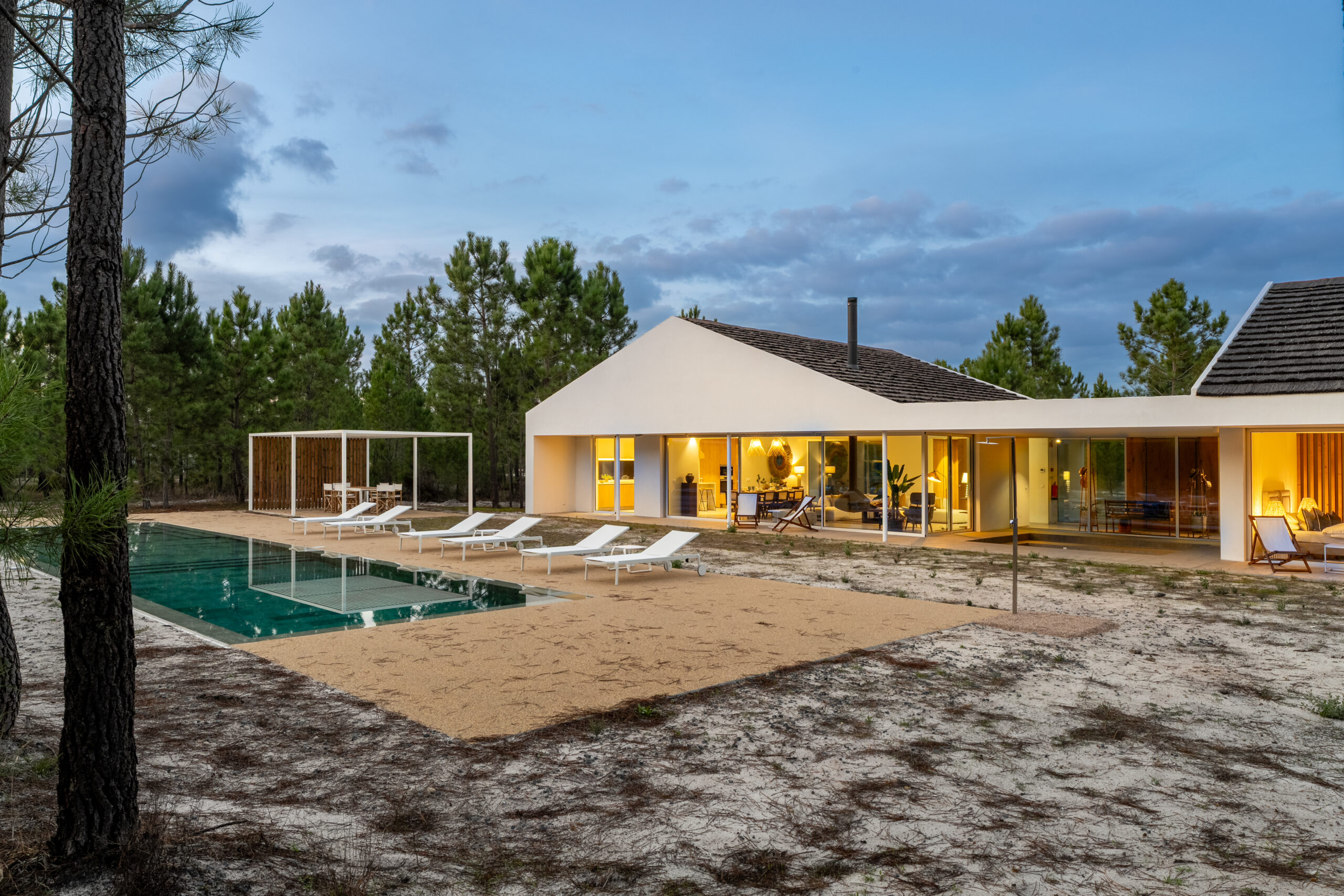 A modern house with large windows is brightly lit at dusk. Featuring a rectangular swimming pool, lounge chairs, and a sandy area, this A-list February 2026 property is bordered by pine trees.