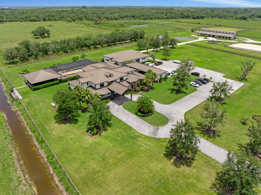 Aerial view of a large modern estate with multiple buildings, winding driveway, neatly mowed lawns, and parked cars—like the perfect retreat for Aquarius season—nestled among open green fields under a blue sky.