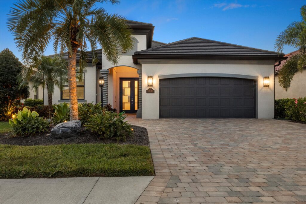 Modern single-story house with light exterior, dark roof, and double garage. Palm trees and landscaping decorate the front yard, while warm lights illuminate the entrance at sunset, perfect for relaxing during Aquarius season.