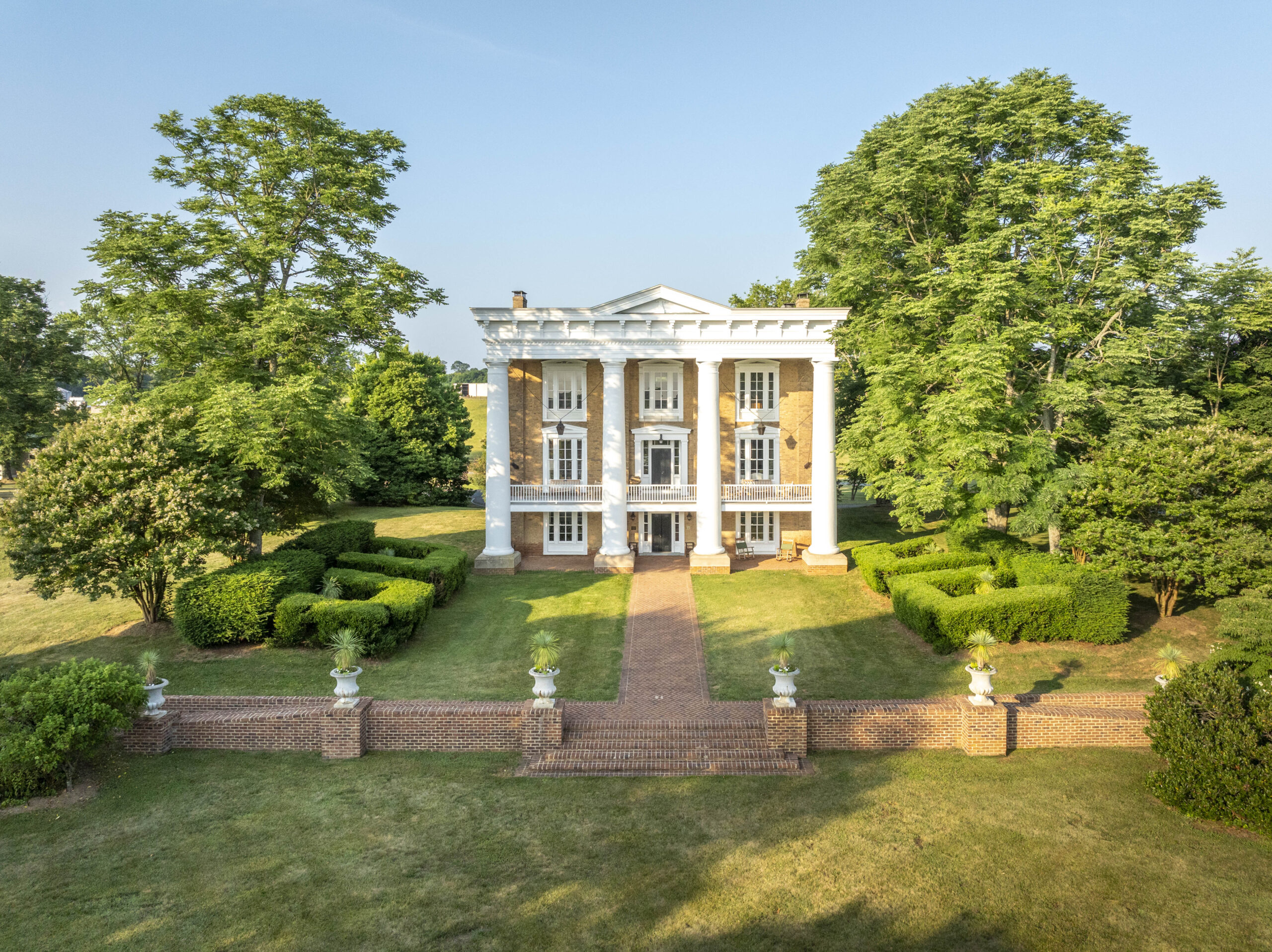 A large, historic, yellow brick mansion with tall white columns and manicured gardens—perfect for an A-list event—stands surrounded by green trees and a brick walkway leading to its grand entrance.