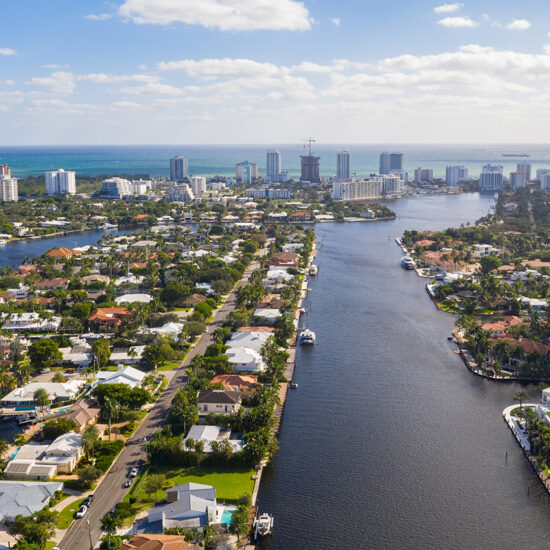 Aerial view of a coastal city showcasing South Florida real estate—waterways lined with houses, boats docked along canals, and tall buildings rising in the background beneath a partly cloudy sky.
