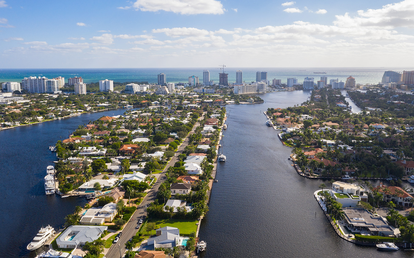 Aerial view of a coastal city showcasing South Florida real estate—waterways lined with houses, boats docked along canals, and tall buildings rising in the background beneath a partly cloudy sky.