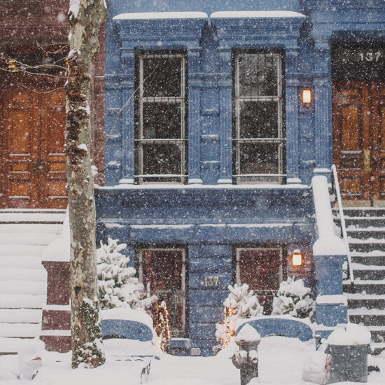 Snow falls heavily on two adjacent townhouse entrances—one brown, one blue—in this Brooklyn condo scene. Steps, doors, and trees are blanketed in fresh snow, while warm lights near the blue door add a cozy contrast to the wintry setting.