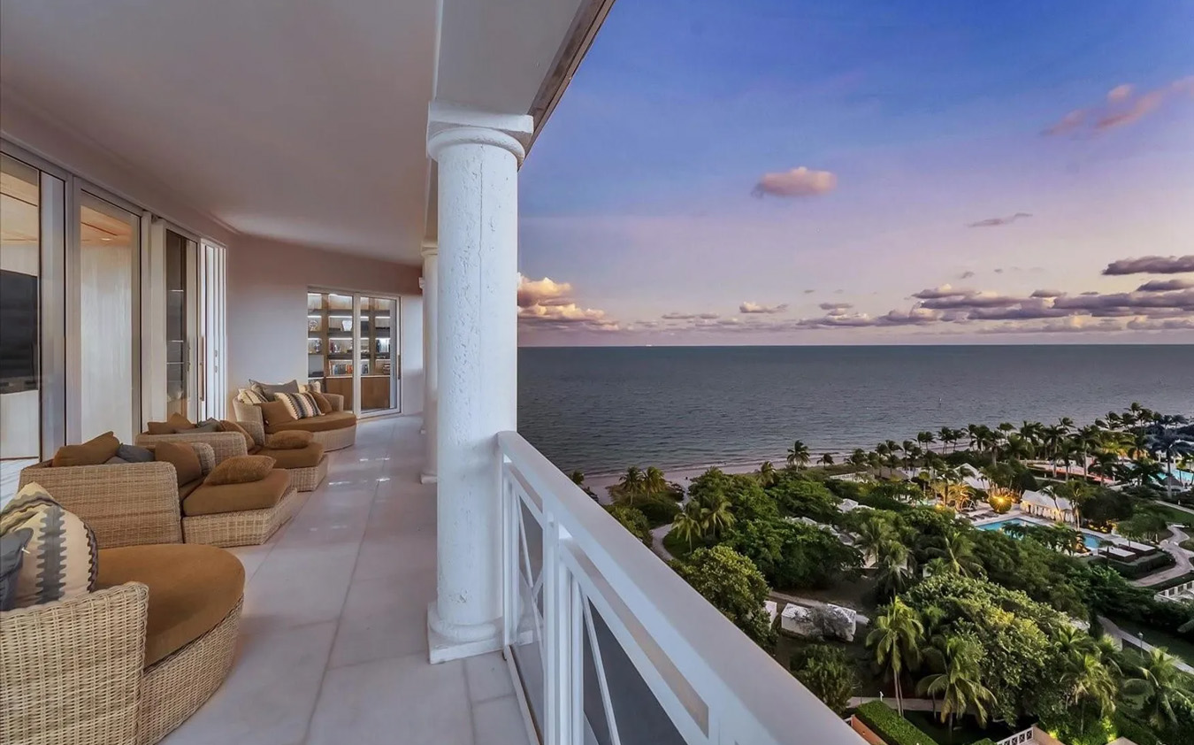 A spacious balcony with wicker chairs and cushions overlooks a lush green landscape, a swimming pool, and the ocean under a colorful sunset sky with scattered clouds.