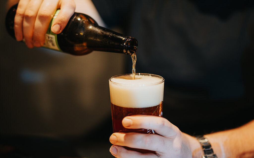 A person pours beer from a brown bottle into a glass, creating a frothy head. The individual, possibly taking a break from holiday decorating, wears a dark shirt and watch, while the background is softly blurred.