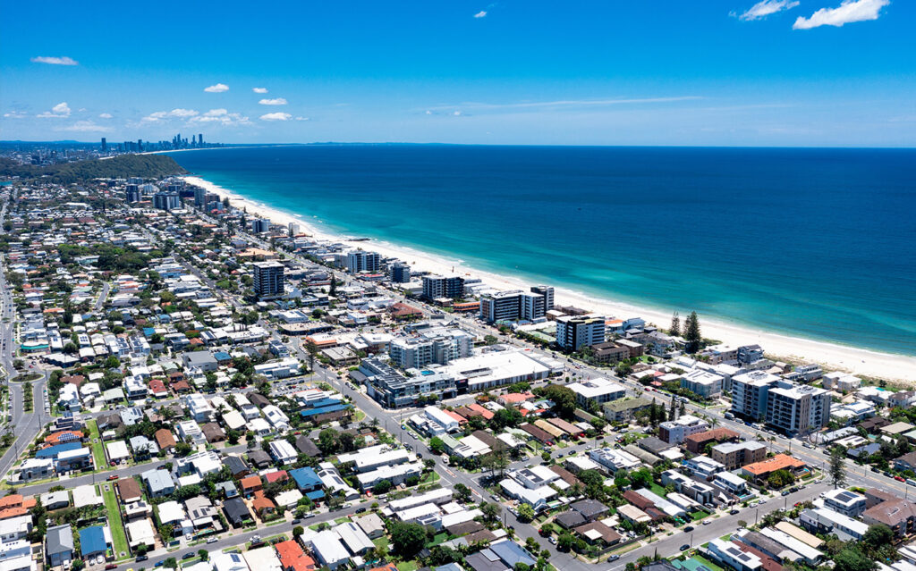 Aerial view of a coastal city with white sandy beaches, turquoise ocean water, and rows of buildings and houses along the shoreline under a clear blue sky—showcasing stunning South Florida Real Estate.