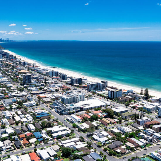 Aerial view of a coastal city with white sandy beaches, turquoise ocean water, and rows of buildings and houses along the shoreline under a clear blue sky—showcasing stunning South Florida Real Estate.