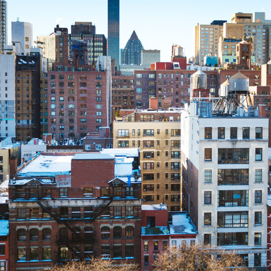A cityscape shows several mid-rise and high-rise buildings with brick exteriors, classic water towers on rooftops, and sunlight illuminating this vibrant NYC residential rental scene under a clear sky.