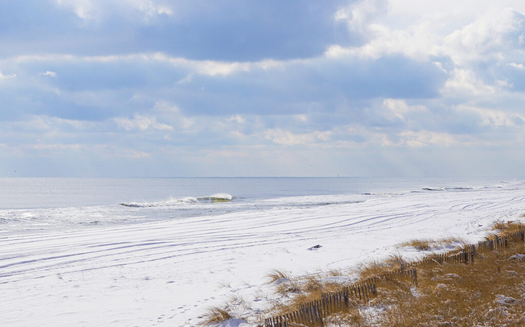 sm_winter_q4 A quiet beach scene in winter, with snow covering the sand, gentle waves in the ocean, and a cloudy sky. Brown grass and a wooden fence line the edge of the snowy beach—a serene view often cherished by East End Real Estate seekers.