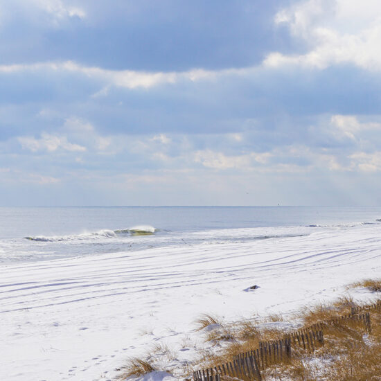 A quiet beach scene in winter, with snow covering the sand, gentle waves in the ocean, and a cloudy sky. Brown grass and a wooden fence line the edge of the snowy beach—a serene view often cherished by East End Real Estate seekers.