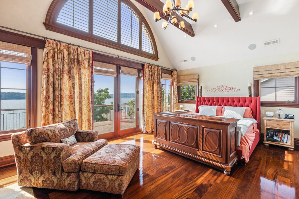 33 Livingston Avenue Spacious, sunlit primary suite with hardwood floors, an ornate bed featuring a red tufted headboard, patterned armchair, large windows with drapes, and a serene view of water and trees outside.