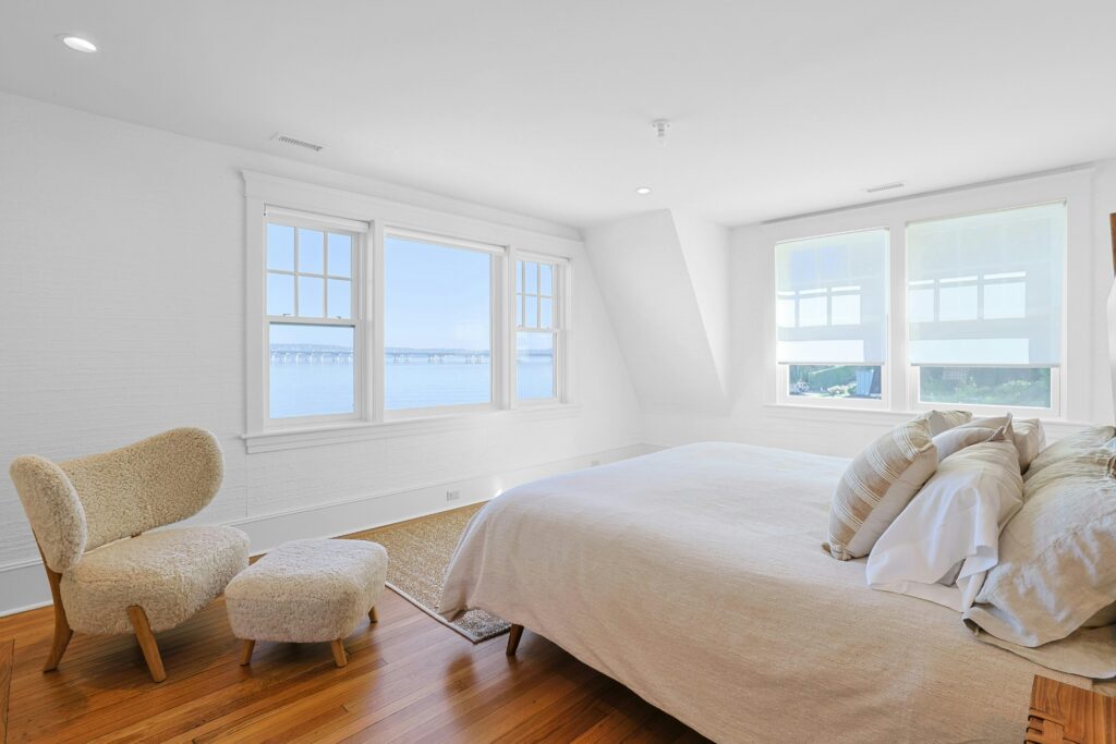 1 Washington Avenue Bright primary suite bedroom with large windows showcasing a water view, a cozy beige bed with several pillows, a matching chair and ottoman, and hardwood floors, creating a peaceful and airy atmosphere.