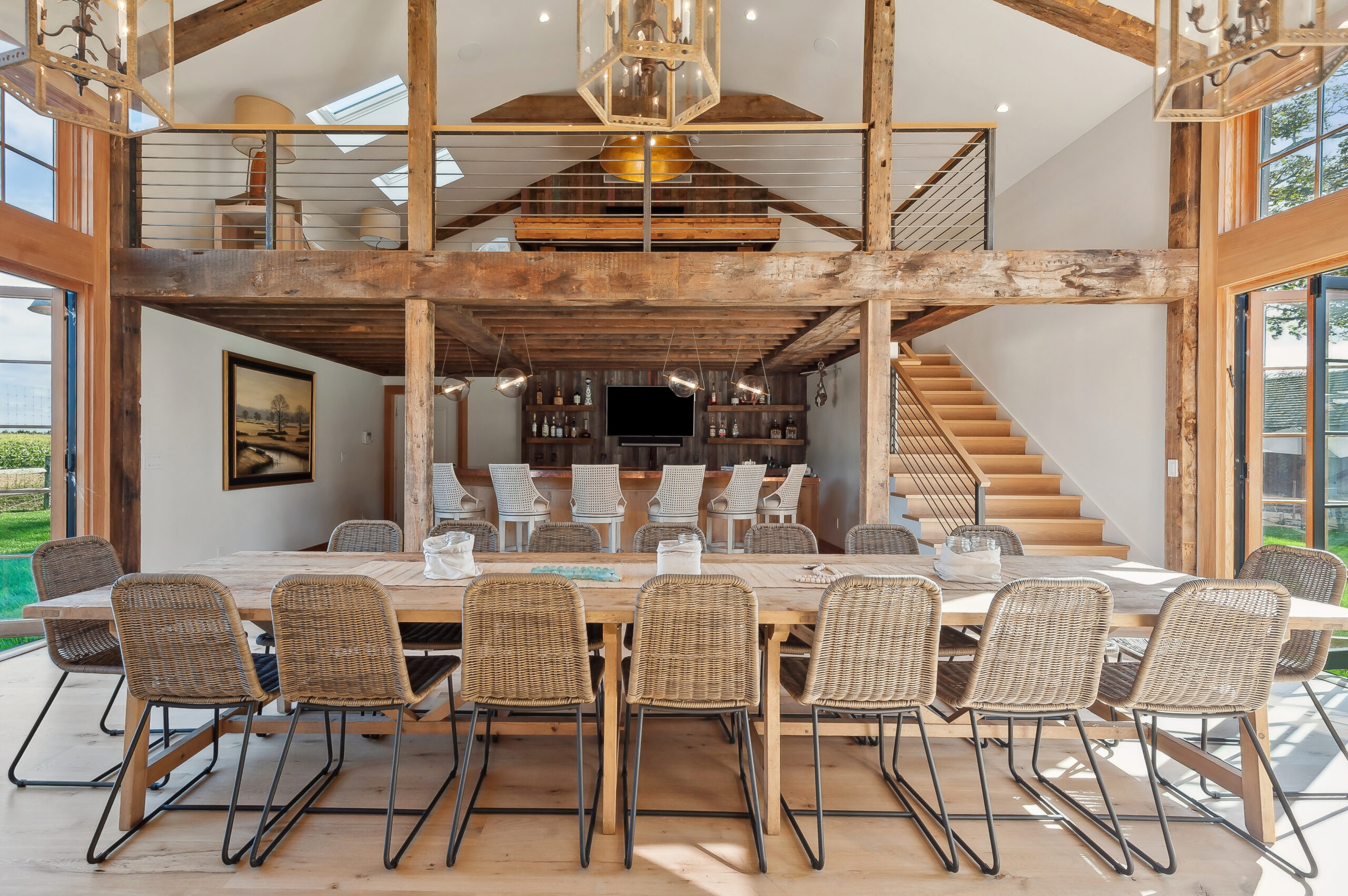 Spacious, rustic dining area with a long wooden table and wicker chairs, open loft above, wooden beams, and a staircase to the right—perfect for gathering friends to watch the big game at the bar area with stools in the background.