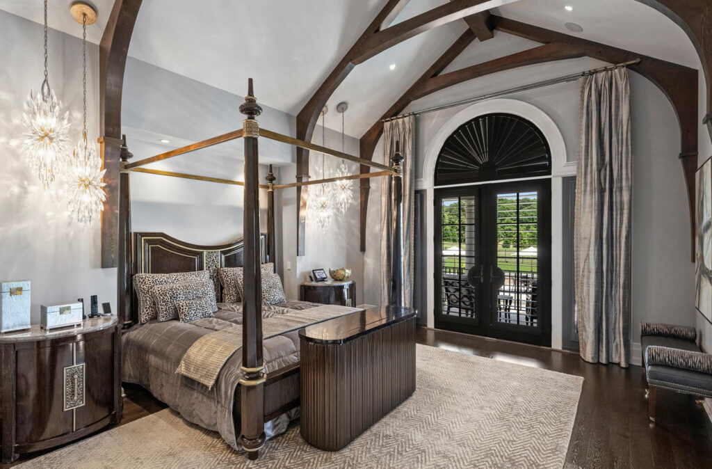 18308 Shearer Road A luxurious primary suite with a vaulted ceiling, exposed wooden beams, a four-poster bed with patterned bedding, two hanging crystal chandeliers, large arched windows with curtains, and elegant dark wood furniture.