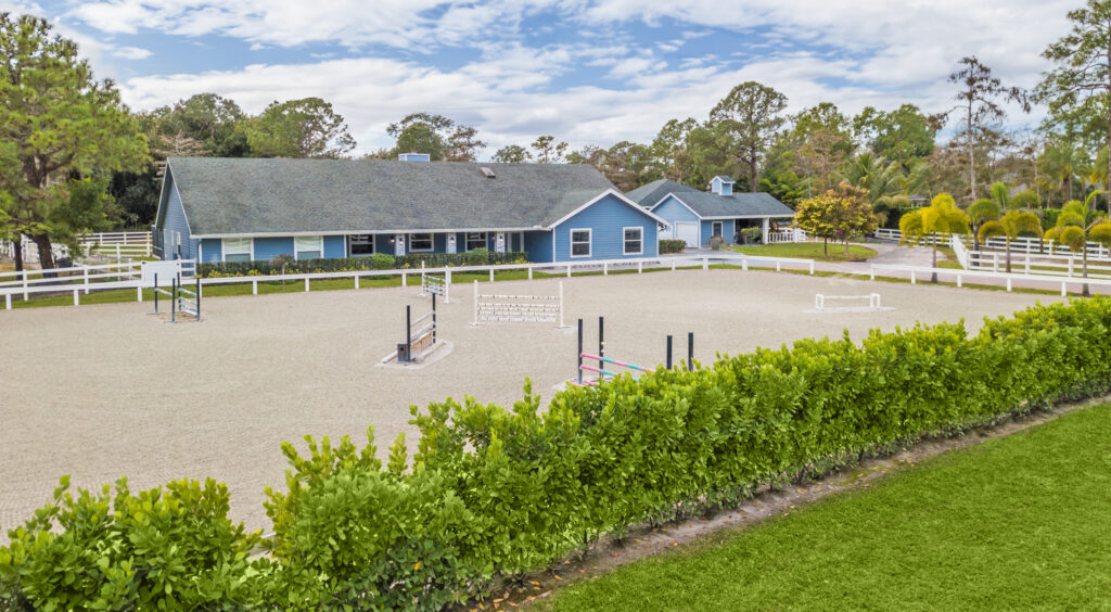 A large, sandy equestrian arena with various jumping obstacles, surrounded by green hedges and white fences, sits in front of a blue ranch-style house reminiscent of classic ski houses, with a barn and trees under a partly cloudy sky.