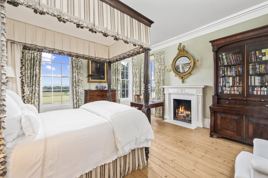 199 Eichybush Road Elegant primary suite featuring a four-poster bed with white bedding, a lit fireplace, antique wooden furniture, gold-framed mirror, bookshelves, and large windows overlooking a green landscape.