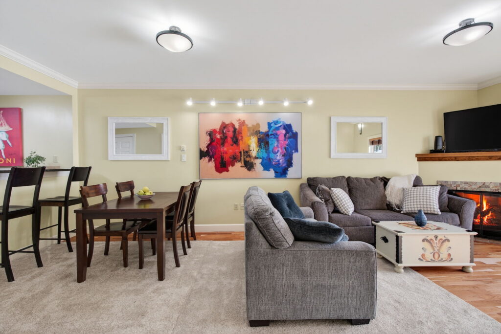 A modern living and dining area, just steps from the primary suite, features a gray sectional sofa, a wooden dining table with six chairs, a fireplace, colorful abstract wall art, and two mirrors on a pale yellow wall.