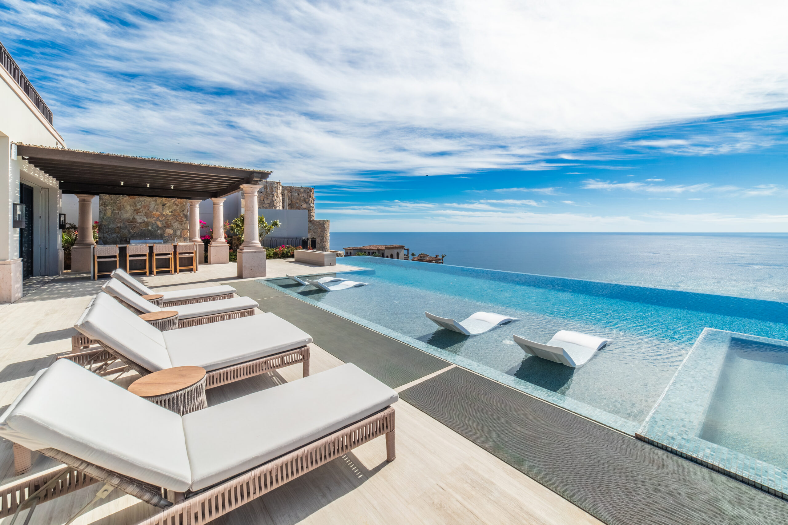 Luxurious outdoor pool area with white lounge chairs and in-pool loungers overlooking a calm, blue ocean under a bright sky, featured on the A-List: March 2026, with a covered dining area to the side.