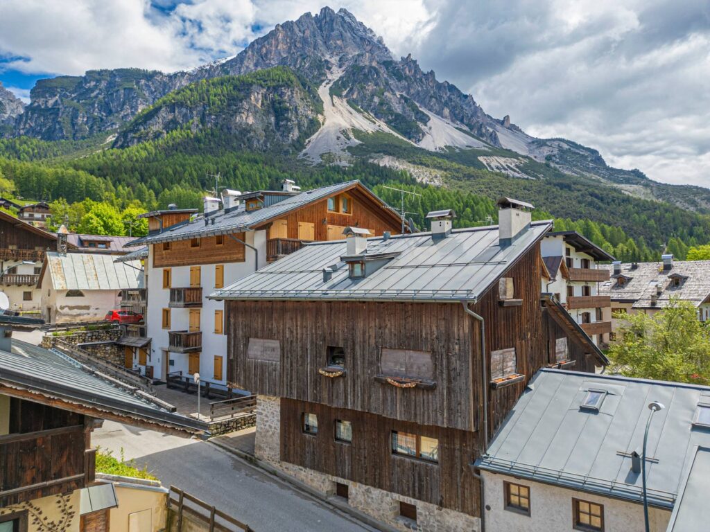 A mountain village with wooden alpine houses, many featuring a cozy primary suite, sloped metal roofs, and a scenic backdrop of tall, forested mountains under a partly cloudy sky.