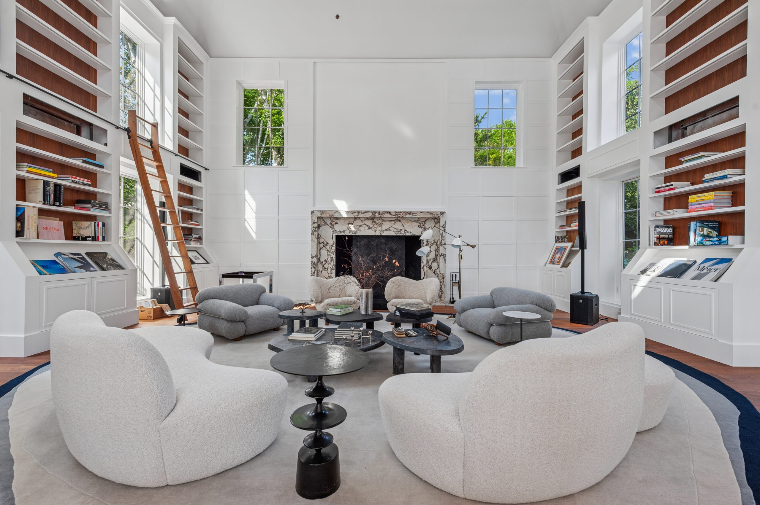 Spacious modern living room with tall bookshelves, rolling ladder, large white armchairs, circular black coffee tables, marble fireplace, and tall windows letting in natural light—a-list: March 2026 style and sophistication.