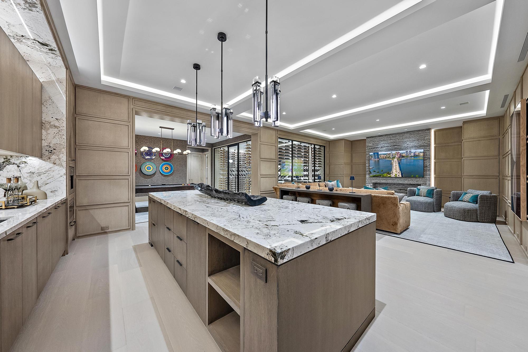 A modern, spacious kitchen with a large marble island, light wood cabinets, and pendant lighting—perfect for prepping snacks for the big game—and an open view into a living area with contemporary art and comfortable seating.