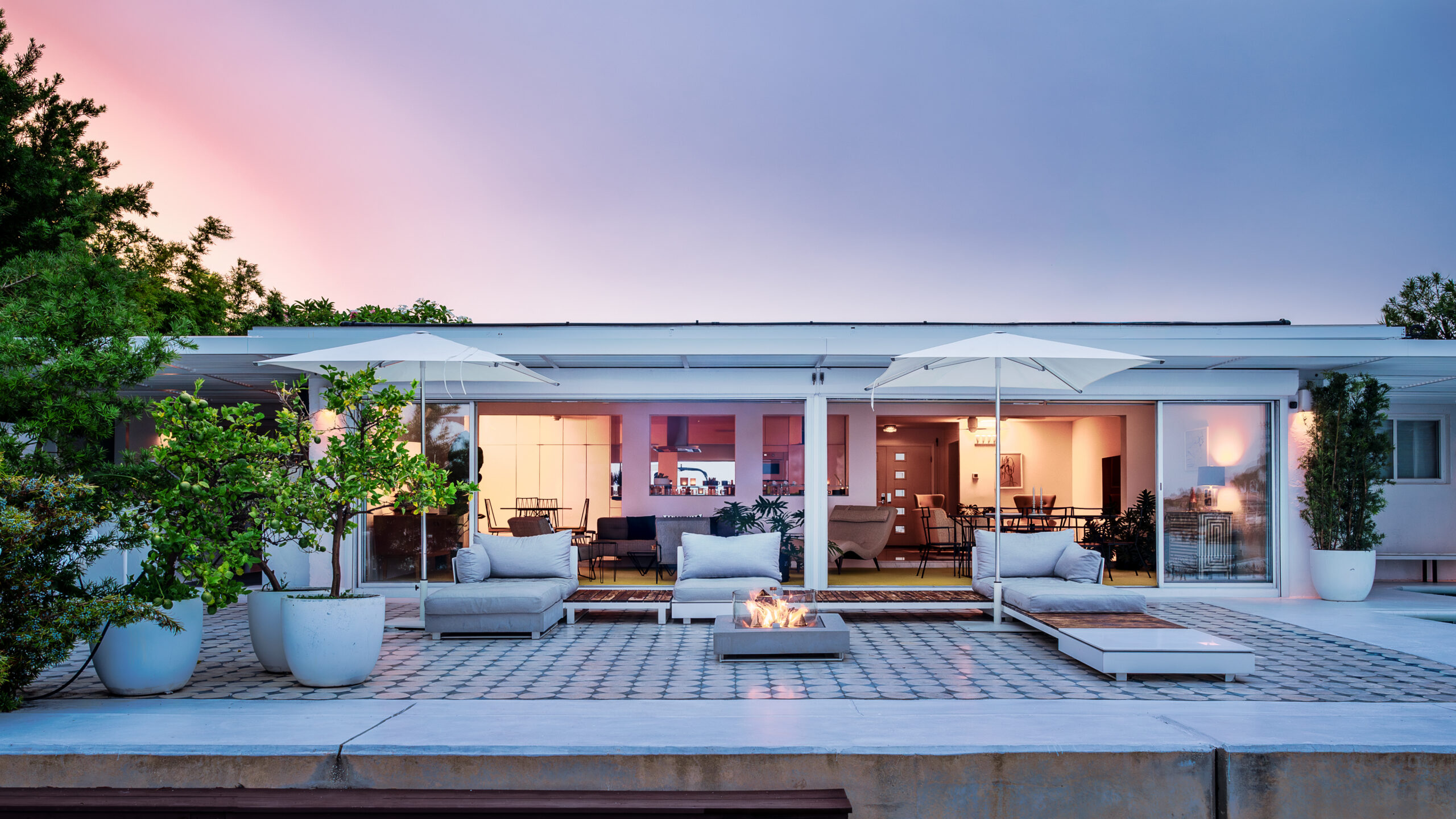 A modern patio with cushioned seating, white umbrellas, potted plants, and a rectangular fire pit in the center, featured in the a-list: February 2026 edition, shown at dusk with warm lighting inside a spacious, open house in the background.