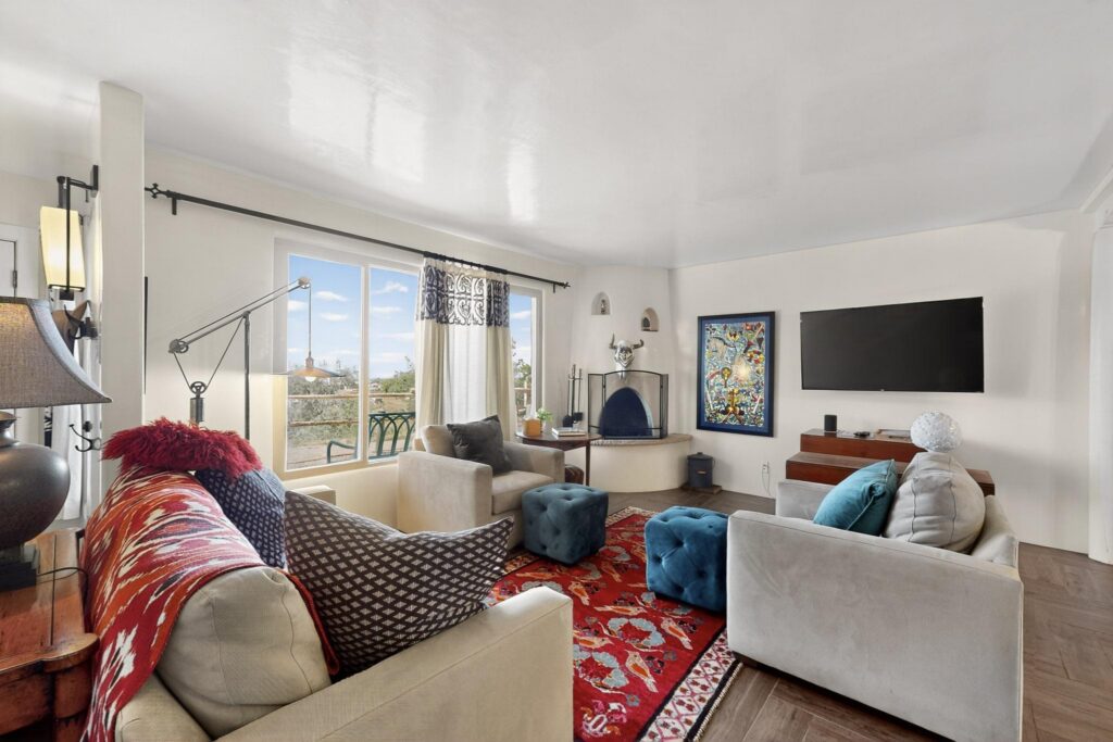 A cozy living room with large windows, modern furniture, a red patterned rug, a blue ottoman, a wall-mounted TV, and a fireplace. Colorful decor and natural light fill the space adjacent to the primary suite.