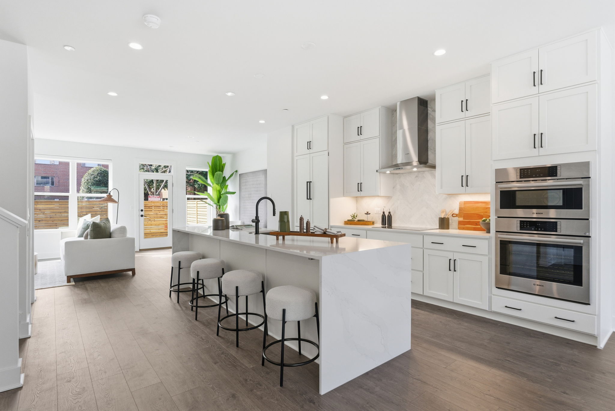 Modern, bright kitchen with white cabinets, marble island, four cushioned bar stools, double wall ovens, stainless steel appliances, wood flooring, and an open layout—a-list style for February 2026—leading to a sunlit living area with large windows.