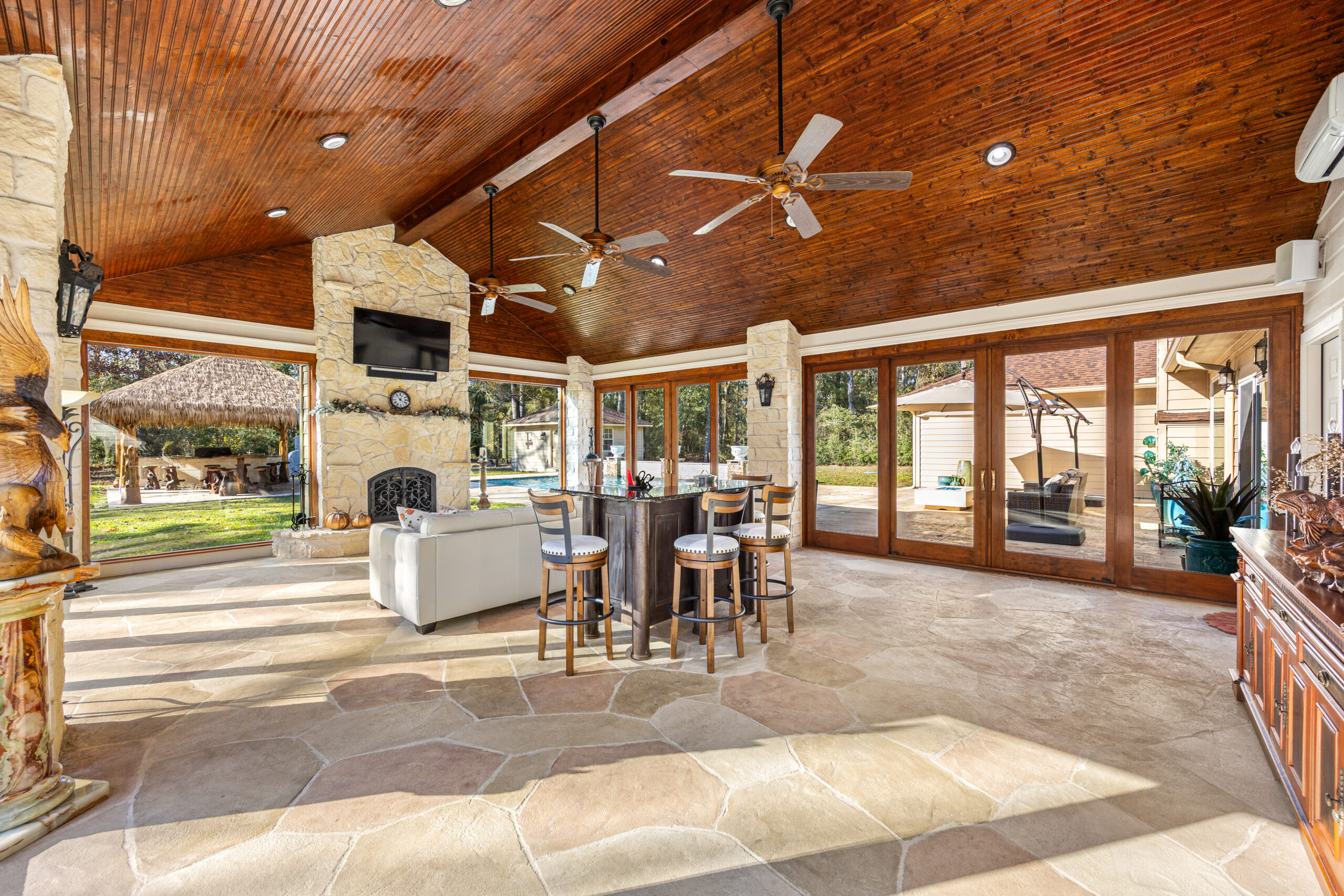 Spacious covered patio with a wooden vaulted ceiling, ceiling fans, stone flooring, bar area with stools—perfect for watching the big game—lounge seating, fireplace, TV, and large glass doors opening to a backyard with pool and gazebo.