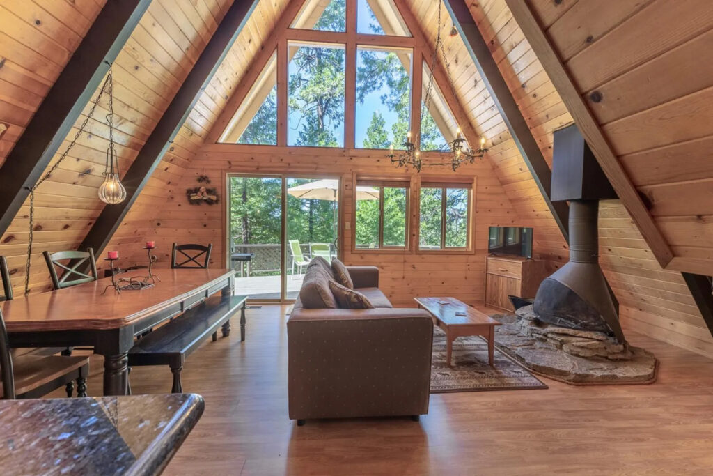 A cozy A-frame cabin interior with wood-paneled walls, large windows, a sofa, a dining table, a wood-burning stove on a stone hearth, and a view of trees through glass doors leading to a deck—plus easy access to the primary suite for comfort and convenience.