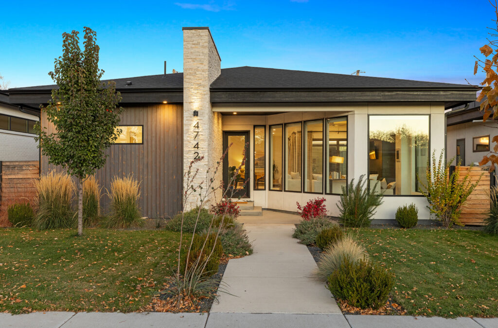 A modern single-story house with large windows, a mix of wood and white exterior, a stone chimney, and a well-maintained front yard.