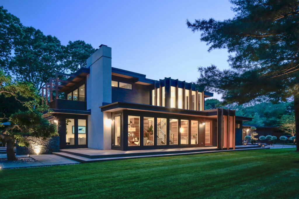 Modern two-story house with large glass windows and warm indoor lighting, surrounded by a green lawn and trees at dusk. Contemporary architectural design features clean lines—a tranquil retreat perfect for reflecting during Pisces season.