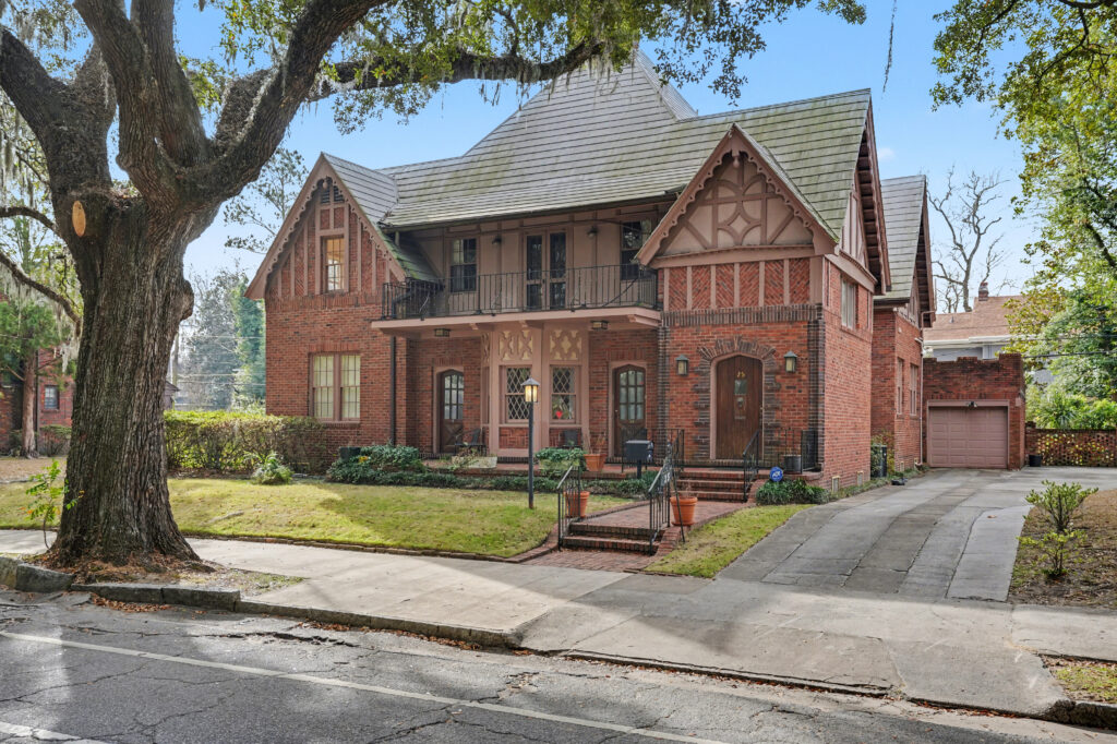 A two-story red brick Tudor-style house with decorative timbering and a steep roof sits under a large tree, its well-kept lawn glowing in pisces season. Front steps lead up past a small balcony and driveway to the detached garage.