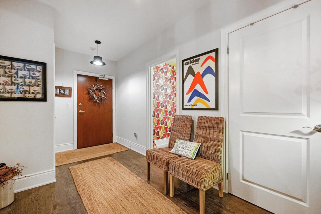 A hallway with a wooden floor and a brown door, a wreath hanging on the door, two wicker chairs with small cushions, abstract wall art, a woven rug, and the vibrant primary suite visible through an open doorway.