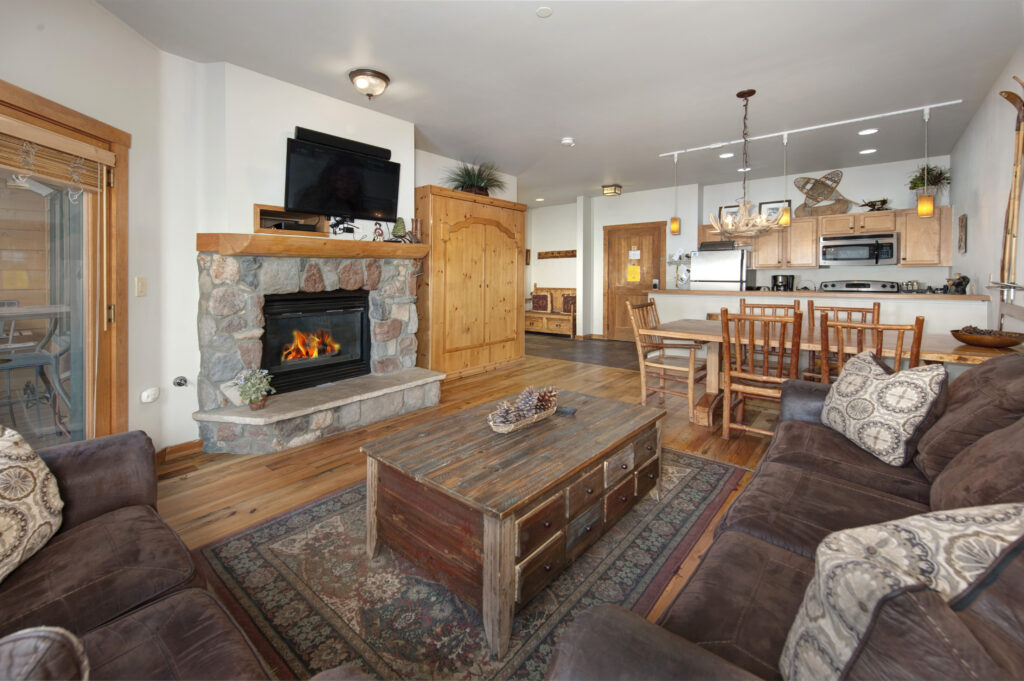 Cozy living room with stone fireplace, brown sofas, rustic wooden coffee table, and a flat-screen TV. An open kitchen and dining area with wooden chairs are visible in the background, just steps away from the inviting primary suite.
