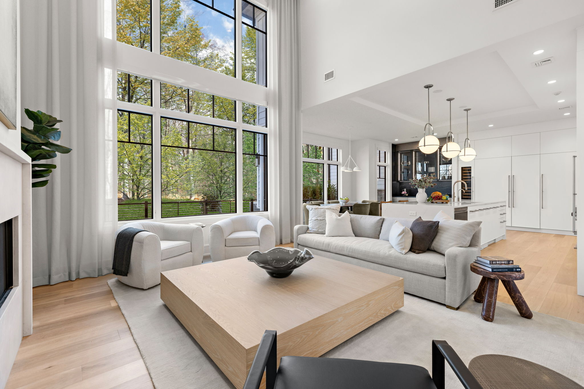 Modern living room with tall windows, neutral-toned furniture, light wood floors, and an open layout connecting to a kitchen with white cabinetry and pendant lights above an island—a-list: March 2026 style. Trees are visible outside.