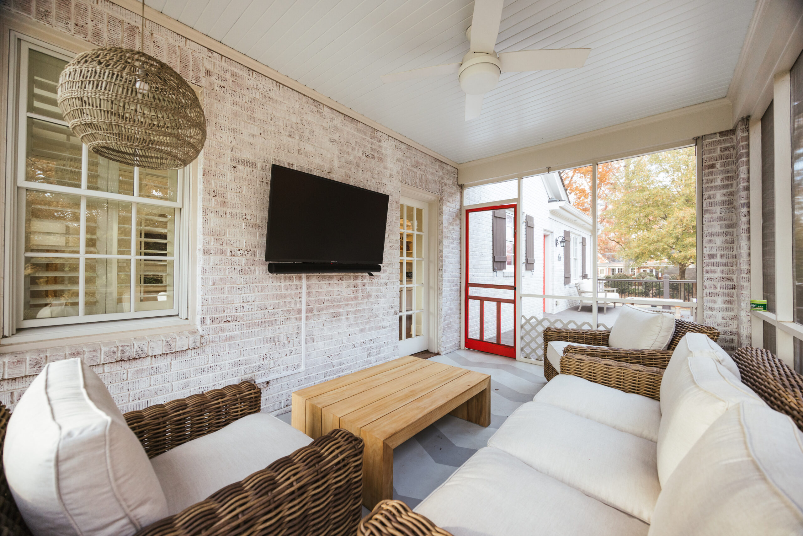 A cozy screened porch with wicker furniture, white cushions, a wooden coffee table, a TV perfect for the big game mounted on a whitewashed brick wall, a ceiling fan, and a red door leading outside.