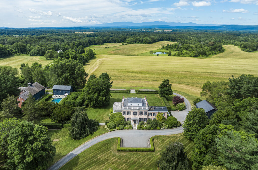 Aerial view of a large estate with a grand house, outbuildings, trees, and a circular driveway—an idyllic retreat reflecting the calm of Pisces season—surrounded by expansive green fields and distant mountains under a partly cloudy sky.
