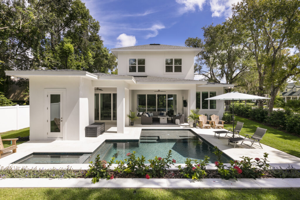 Modern two-story white house with large windows, a patio seating area, and a swimming pool surrounded by greenery and flowers.