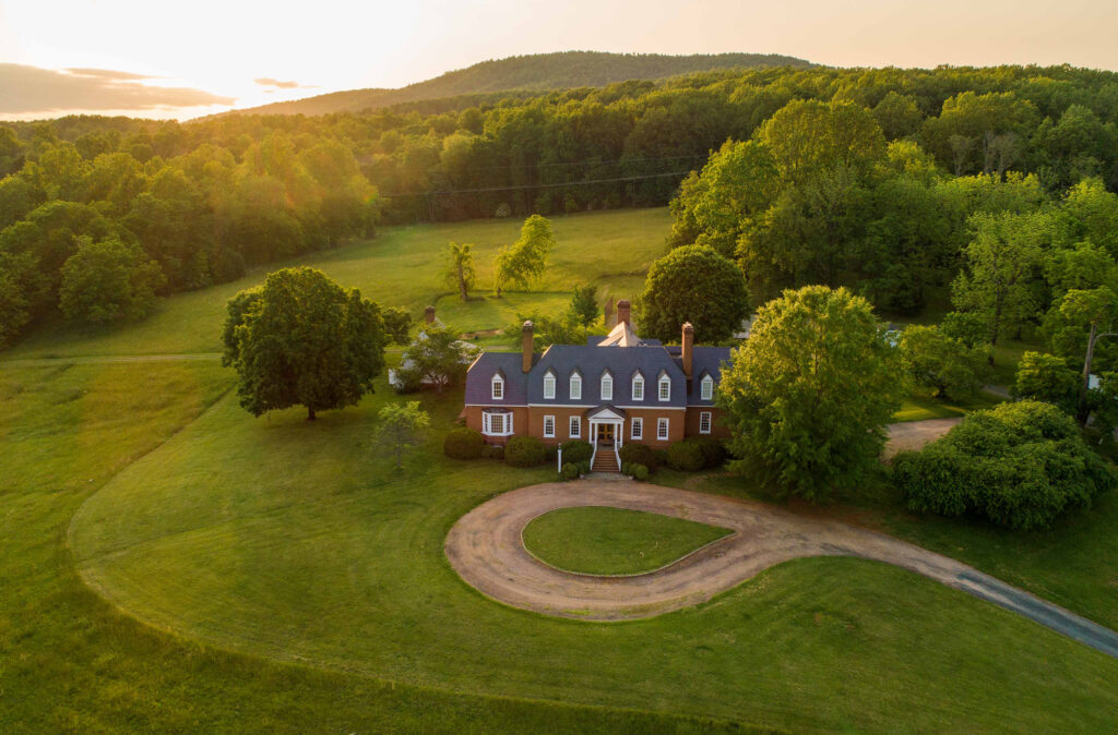 A large brick house with white trim and multiple chimneys sits surrounded by green lawns, trees, and a circular driveway—an inviting retreat to reflect during Pisces season, nestled against wooded hills and sunlight in the background.