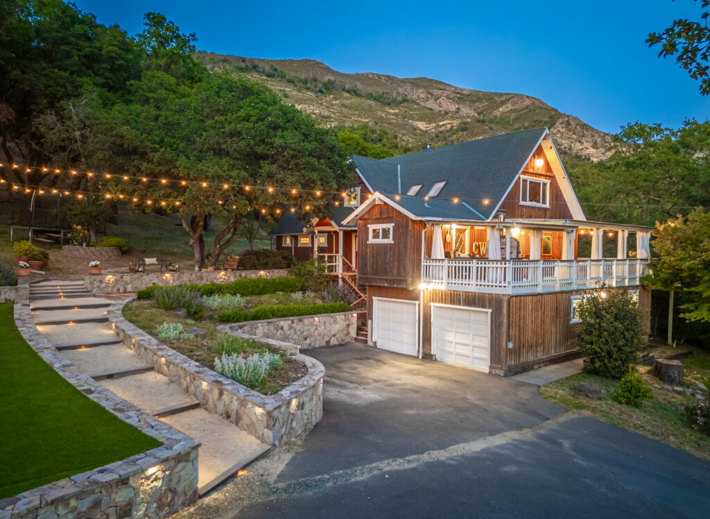 A rustic multi-level house with a wraparound porch and string lights glows at dusk, nestled against a hillside and surrounded by trees, stone pathways, and manicured landscaping—an ideal retreat for reflection during Pisces season.