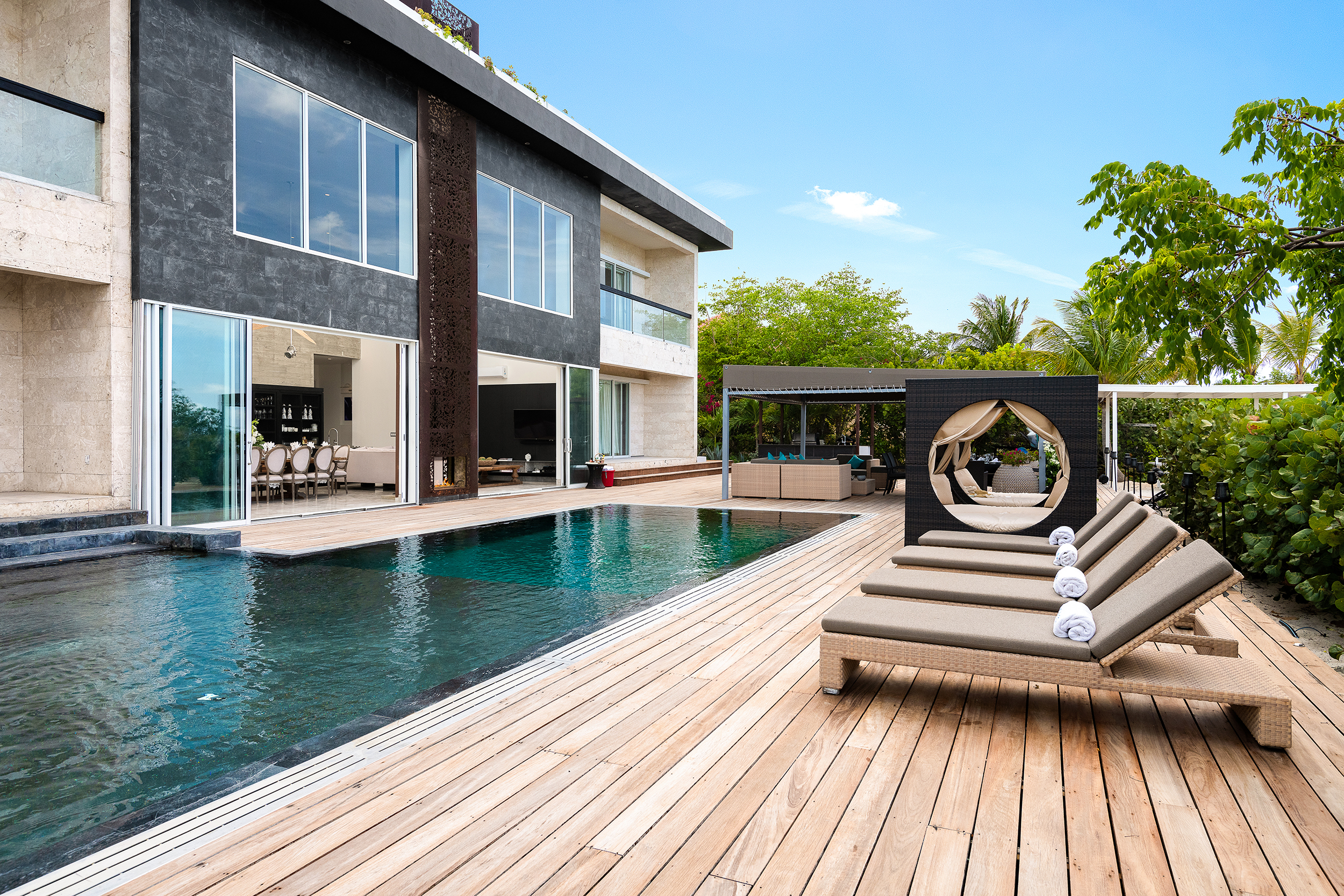 Modern luxury villa with a rectangular pool, wooden deck, sun loungers with rolled towels, and open lounge area. Large windows invite nature in—an A-list retreat for March 2026, surrounded by trees under a clear blue sky.