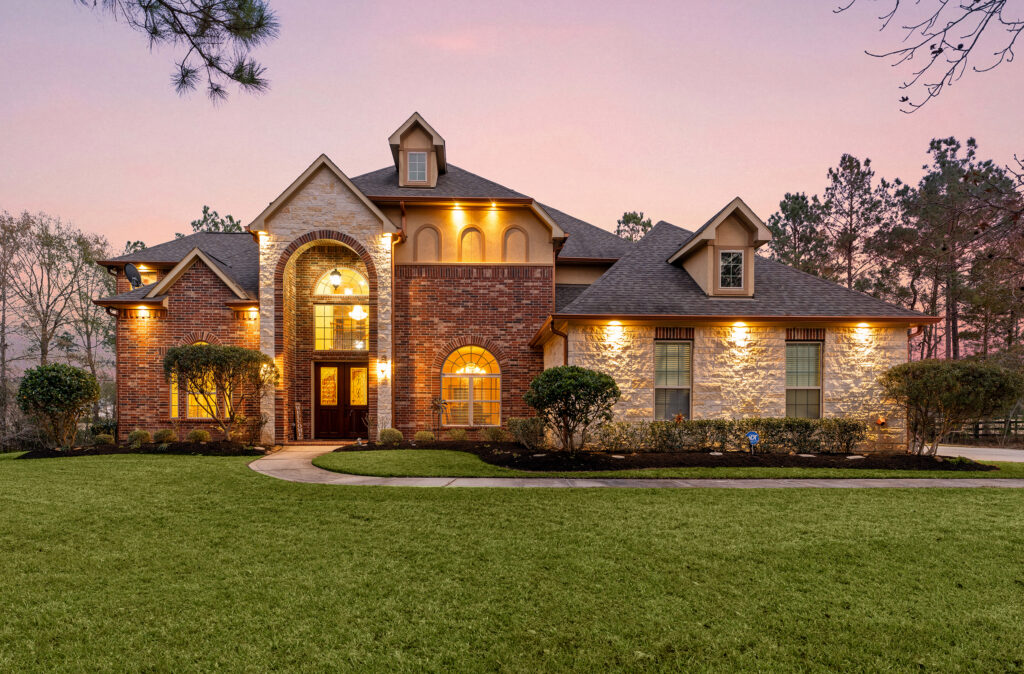 Two-story brick and stone house with arched windows and a well-lit entrance, surrounded by neatly trimmed bushes and a large green lawn at sunset.