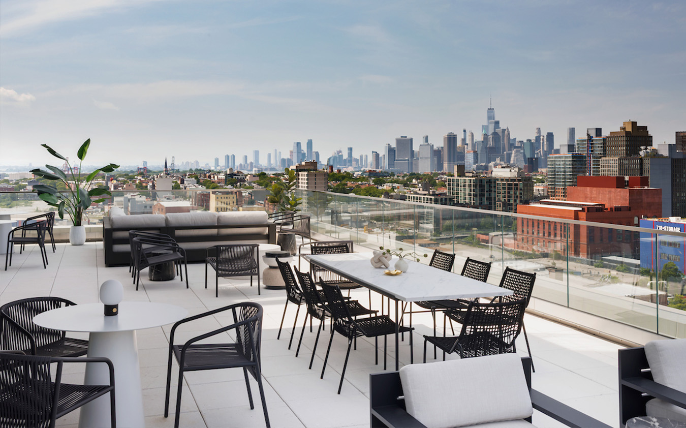 Modern rooftop terrace with black and white outdoor furniture, a dining area, lounge seating, potted plants, and glass railing overlooking a city skyline under a partly cloudy sky.