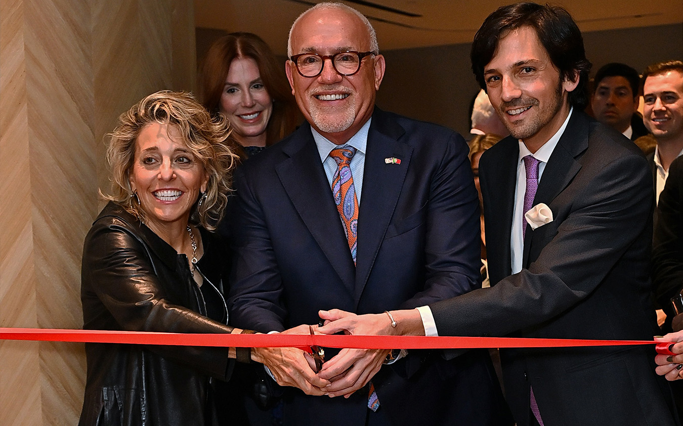 Three people smile and hold scissors together as they cut a red ribbon at an indoor event, with several others standing in the background, signifying a ribbon-cutting ceremony.