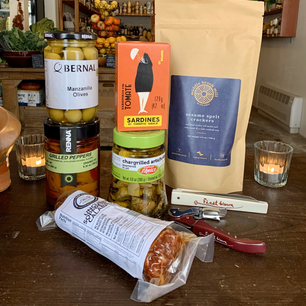 A selection of gourmet foods on a table, including olives, grilled peppers, artichokes, sardines, sesame spelt crackers, cured meat, and a can opener—perfect for holiday decorating with two lit candles in the background.