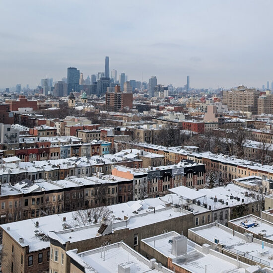 A cityscape view of Brooklyn, New York, in winter, with snow covering rooftops and streets, and a glimpse of the Manhattan luxury sales district and tall buildings visible in the distance under a cloudy sky.