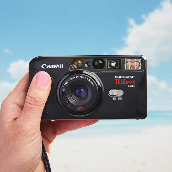 A hand holding a Canon Sure Shot Telemax film camera against a blurred beach background with blue sky, white sand, and turquoise water—capturing moments as memorable as holiday decorating.
